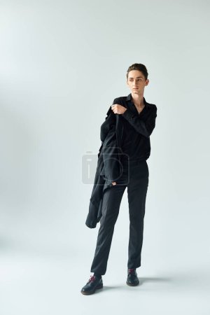 A young queer person, exuding pride, strikes a pose in a studio wearing a black coat and pants on a grey background.