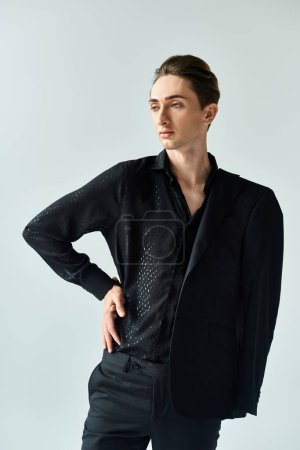 Un joven queer posa en un elegante traje negro contra un fondo gris del estudio, exudando confianza y orgullo.