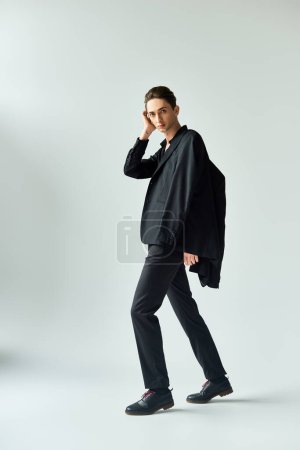 A young queer person strikes a pose in a studio, donning a stylish black jacket and pants, against a grey background.