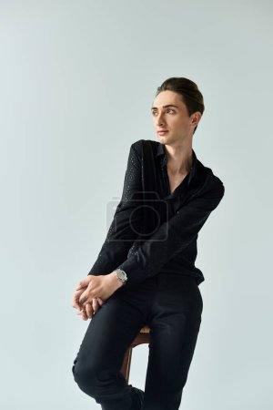A stylish young queer person, representing the LGBTQ+ community, sits on a black stool in a studio against a grey backdrop.