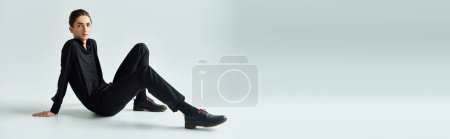 Téléchargez les photos : Un jeune queer en tenue noire, assis gracieusement sur le sol dans un studio avec un fond gris. - en image libre de droit