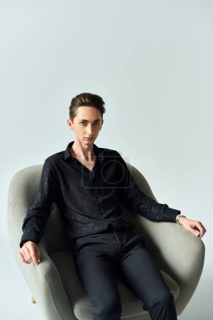 Ein junger queerer Mann posiert selbstbewusst in einem schwarzen Hemd, während er auf einem Stuhl vor grauem Studiohintergrund sitzt.