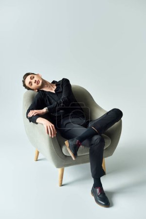 Ein junger queerer Mann lehnt sich anmutig auf einem eleganten grauen Stuhl vor minimalistischem Hintergrund zurück.