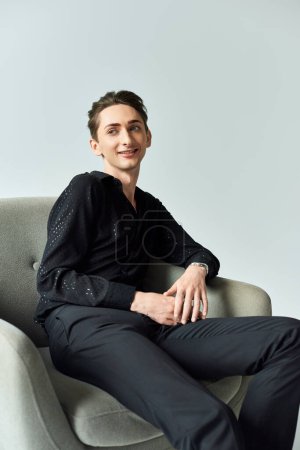 Ein junger queerer Mann sitzt selbstbewusst in einem Stuhl, trägt ein schwarzes Hemd und demonstriert Stolz und Stärke in einem Studio-Setting auf grauem Hintergrund..