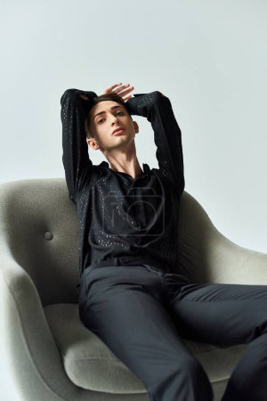 Ein junger queerer Mensch sitzt auf einem Stuhl, die Hände auf dem Kopf, tief in Gedanken, auf einem grauen Studiohintergrund.