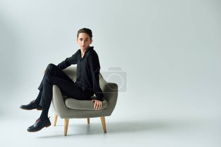 Foto de A young queer person gracefully poses on a chair in a studio against a grey backdrop. - Imagen libre de derechos