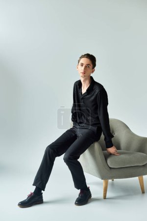 Eine junge queere Person posiert auf einem Stuhl in einem Studio mit grauem Hintergrund.