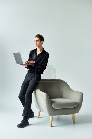 Ein junger queerer Mensch sitzt auf einem Stuhl mit einem Laptop und drückt Kreativität und Inspiration aus, umgeben von einer modernen Atmosphäre.