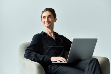 A young queer person on a grey background sits in a chair with a laptop, exuding confidence and pride in their digital presence.