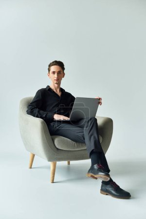 Un joven queer se sienta en una silla con un portátil, profundamente absorto en su trabajo, sobre un fondo gris de estudio.