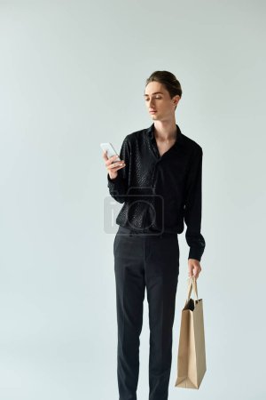 A young queer person holding a shopping bag, looks at his phone against a grey background, showcasing multitasking in urban style.