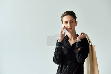 Eine junge queere Person hält Einkaufstüten in der Hand, während sie vor grauem Studiohintergrund telefoniert.