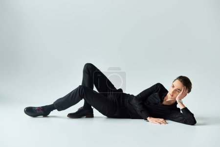 A young queer person elegantly poses in a black suit on a grey studio floor, exuding confidence and pride.