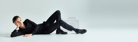 Ein junger queerer Mensch liegt gedankenverloren mit den Händen auf den Hüften auf dem Boden vor grauem Studiohintergrund.