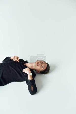 Ein junger queerer Mensch liegt auf dem Bauch, strahlt Ruhe und Introspektion aus, vor einem schroffen weißen Hintergrund.