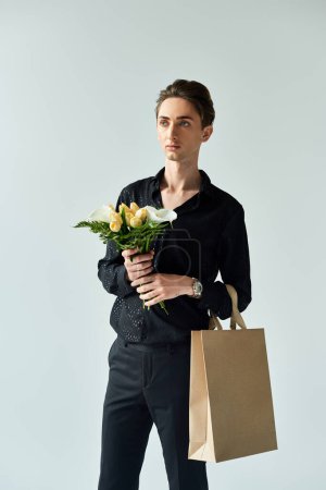 A young queer man elegantly holds a paper bag filled with vibrant flowers, radiating pride in a studio setting.