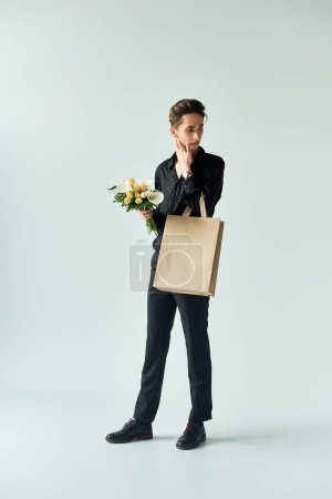 A young queer person strikes a pose holding a shopping bag and a bouquet of flowers on a grey studio background.
