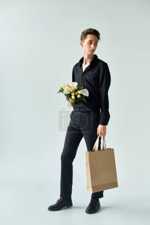A young queer person holding a paper bag filled with colorful flowers against a grey background.