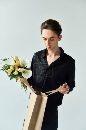 Ein junger queerer Mensch hält stolz eine Papiertüte voller Blumen in einem Atelier vor grauem Hintergrund.