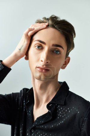 A young queer person strikes a thoughtful pose with his hands resting on his head in a studio setting against a grey background.