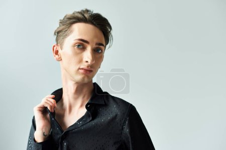 Téléchargez les photos : Un jeune homme en chemise noire pose gracieusement dans un studio sur fond gris, mettant en valeur sa fierté pour la communauté LGBT. - en image libre de droit