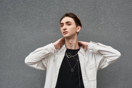 A young queer person confidently poses against a gray wall, showcasing his stylish attire with pride and confidence.