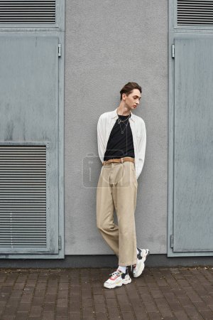Photo for A young queer person in tan pants and a white shirt leans casually against a wall, exuding confidence and style. - Royalty Free Image