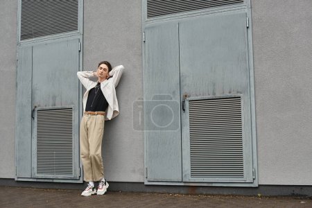 A stylish young queer person leans confidently against a wall, exuding pride and individuality.