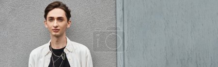 Foto de Un joven queer con un atuendo elegante se para audazmente frente a una pared gris, irradiando orgullo y desafío. - Imagen libre de derechos