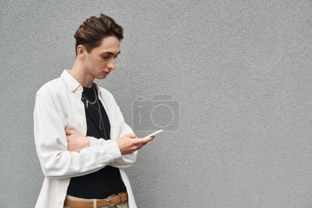 Stilvolle junge queere Person in lässiger Kleidung lehnt an einer Wand und hält ein Handy in der Hand.