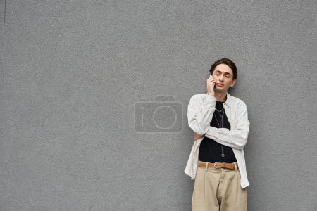 Una persona joven de moda, que se identifica como queer, hablando en un teléfono celular mientras se apoya contra una pared gris.
