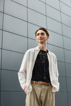 Téléchargez les photos : Un jeune queer vêtu d'une chemise blanche et d'un pantalon bronzé pose en toute confiance devant un immeuble. - en image libre de droit