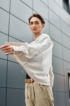 Ein junger queerer Mensch, stilvoll gekleidet in weißem Hemd und brauner Hose, strahlt in seiner Kleidung Zuversicht und Stolz aus..