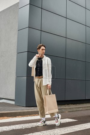 Un joven homosexual vestido con estilo cruza con confianza la calle con una bolsa en la mano.