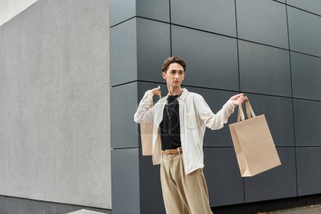 Una persona queer joven con un atuendo elegante sosteniendo bolsas de compras frente a un edificio, abrazando la terapia de venta al por menor.