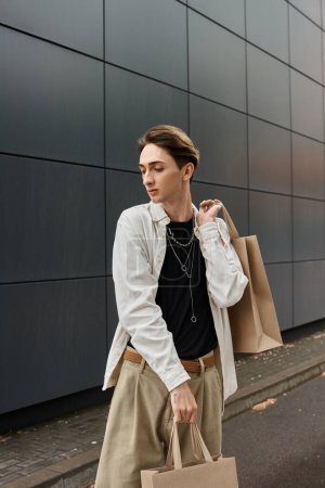 Photo for A young LGBTQ individual in stylish attire carrying shopping bags in front of a modern building. - Royalty Free Image