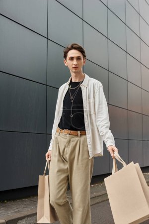 A young queer person in stylish attire, holding shopping bags, stands in front of a modern building.