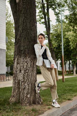 Ein junger queerer Mensch in stilvoller Kleidung lehnt nachdenklich an einem Baum und verkörpert Stolz und Besinnlichkeit.