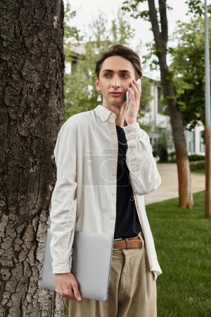 A young, stylishly dressed queer individual standing next to a tree, chatting on his phone in a casual outdoor setting.