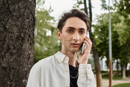 Foto de Un hombre joven, vestido con estilo, conversa en un teléfono celular en un entorno de parque. - Imagen libre de derechos
