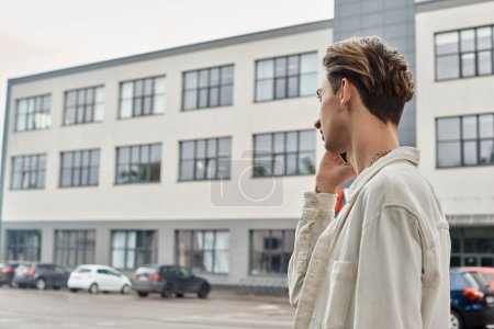 Une jeune personne queer en tenue à la mode parlant sur son téléphone portable devant un immeuble urbain, mettant en valeur la fierté LGBTQ.