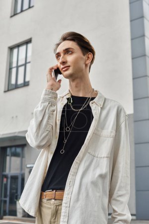 Photo for A young queer individual in stylish attire talks on a cell phone in front of a striking urban building. - Royalty Free Image