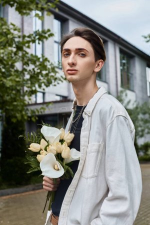 Téléchargez les photos : Un jeune homme élégamment habillé tient fièrement un bouquet de fleurs colorées devant un grand bâtiment. - en image libre de droit