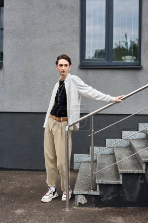 Eine junge queere Person in stilvoller Kleidung steht während einer Stolzereignis selbstbewusst auf den Stufen eines Gebäudes.