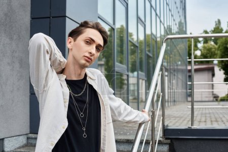 A young man in stylish attire leans on a railing outside a building, exuding confidence and pride in his LGBTQ identity.