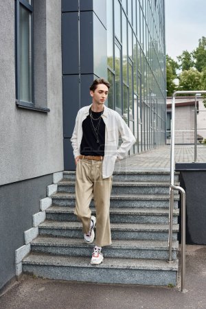 Ein junger queerer Mensch in stilvoller Kleidung posiert selbstbewusst auf den Stufen eines Gebäudes.