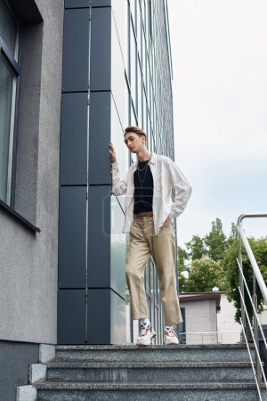Eine junge queere Person in stilvoller Kleidung steht selbstbewusst auf den Stufen eines Gebäudes, das LGBT-Stolz feiert.