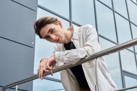 Un joven vibrante, que encarna el orgullo queer, se apoya con confianza en una barandilla frente a un edificio moderno.