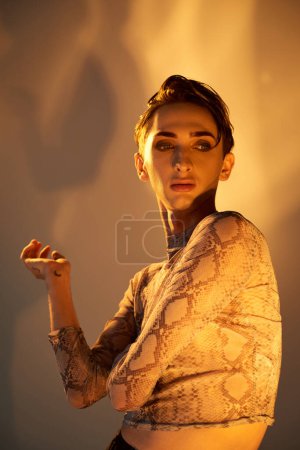 Photo for A young queer person in a snakeskin top strikes a pose in front of a bright light, exuding style and confidence. - Royalty Free Image