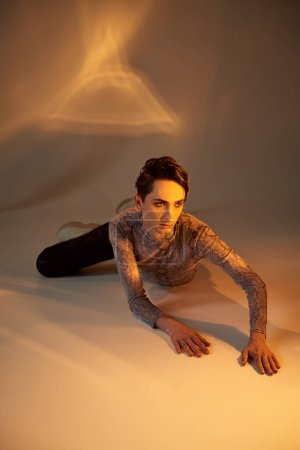 A young queer person in stylish attire lies on the floor, illuminated by a bright light shining directly on him.